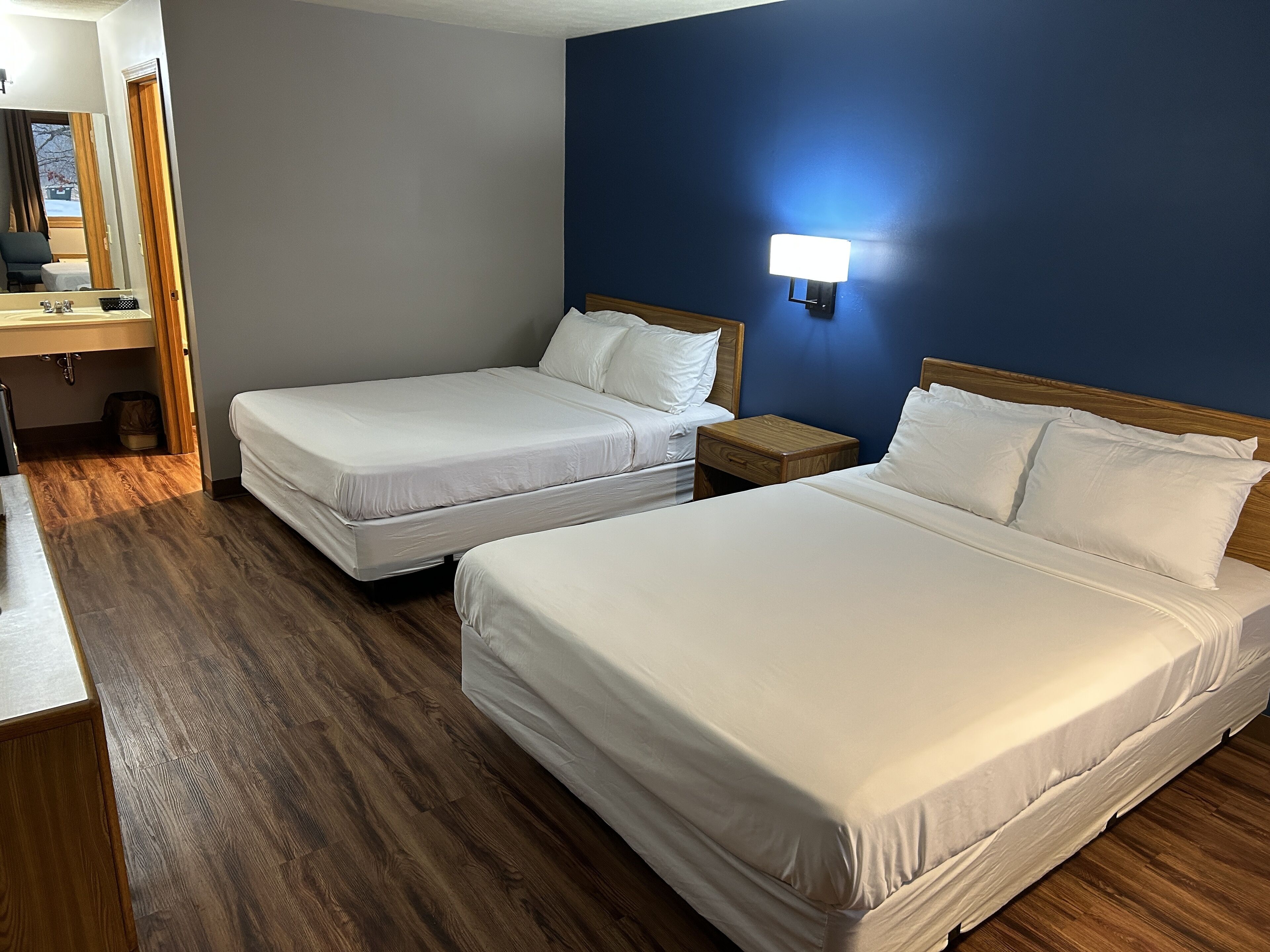 Habitación de hotel con dos camas, suelo de madera, mesita de noche, lámpara y baño con espejo y lavamanos.