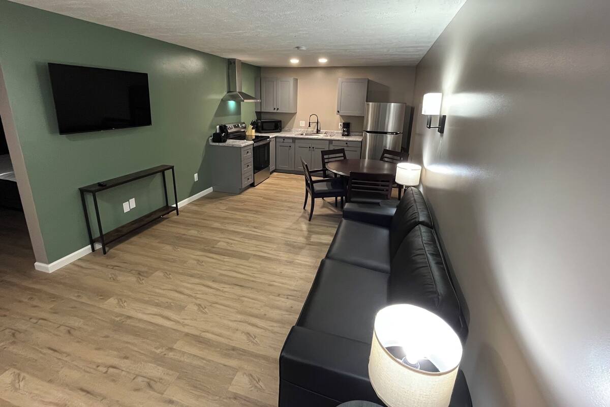 Eine moderne Küche mit Flachbildfernseher, ein Essbereich mit schwarzem Sofa und Tisch, sowie ein Wohnzimmer mit einer Deckenleuchte.