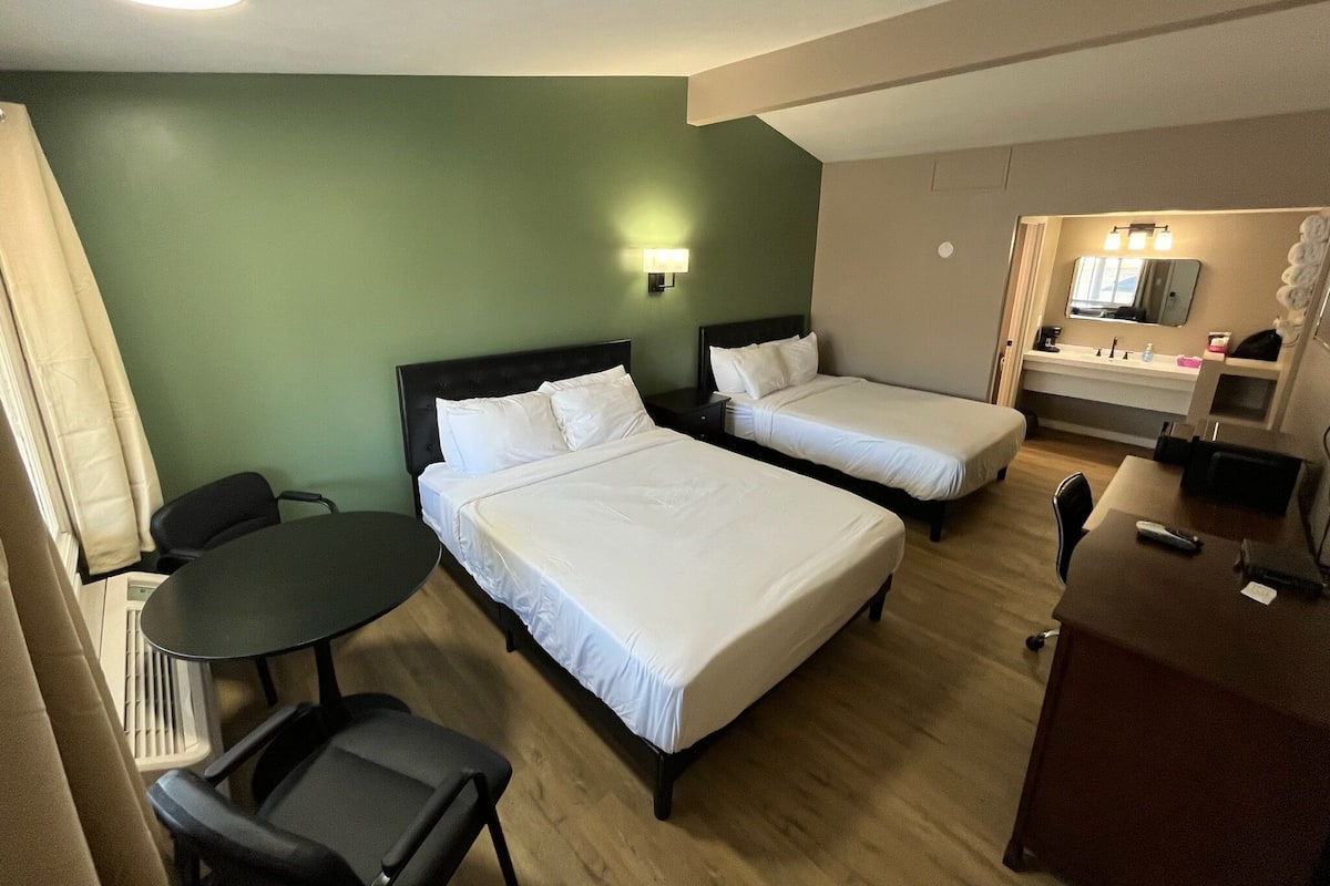 Ein Hotelzimmer mit zwei Betten, einem Schreibtisch, einem Sessel, einem kleinen Tisch, einem Spiegel und einem Waschbecken.