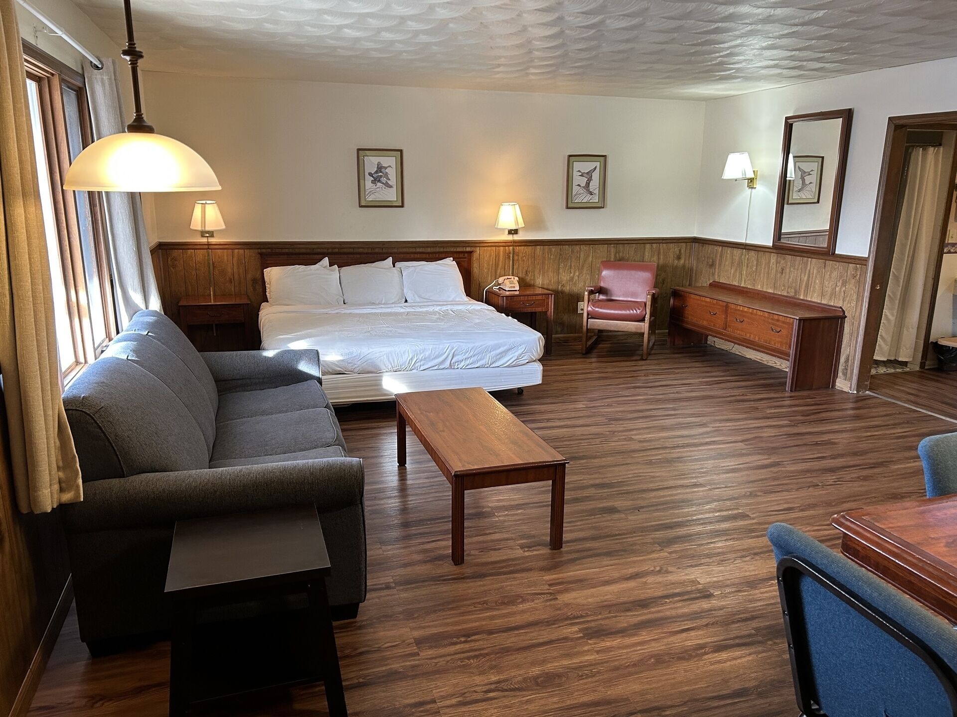 Habitación de hotel con una cama grande, un sofá, una mesa de centro de madera, una silla y un escritorio. Hay dos lámparas de pared y cuadros enmarcados en las paredes.