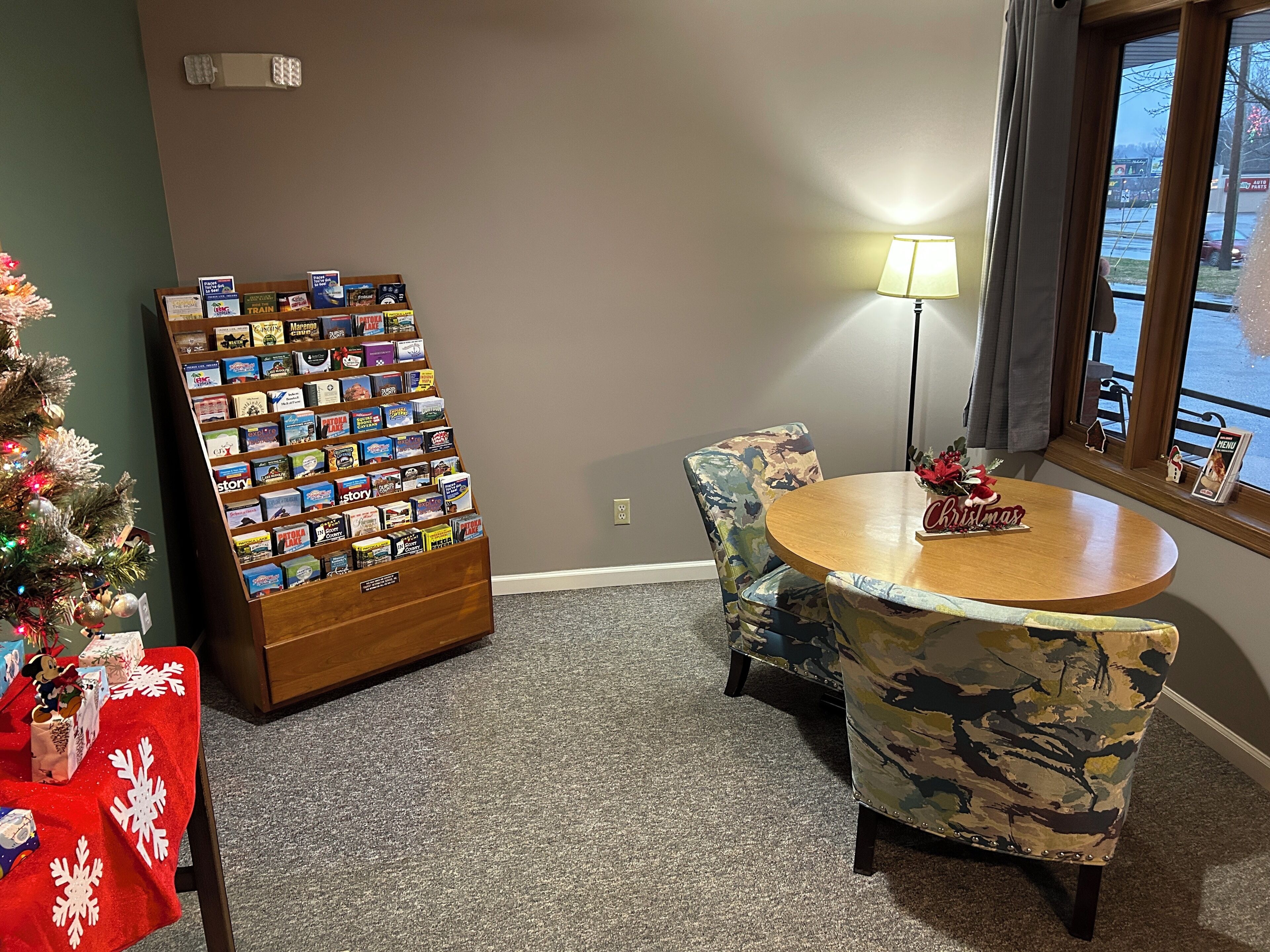 A hotel room with a Christmas tree, a book display, a round table with a 'Christmas' sign, and a view of the outside through a window.