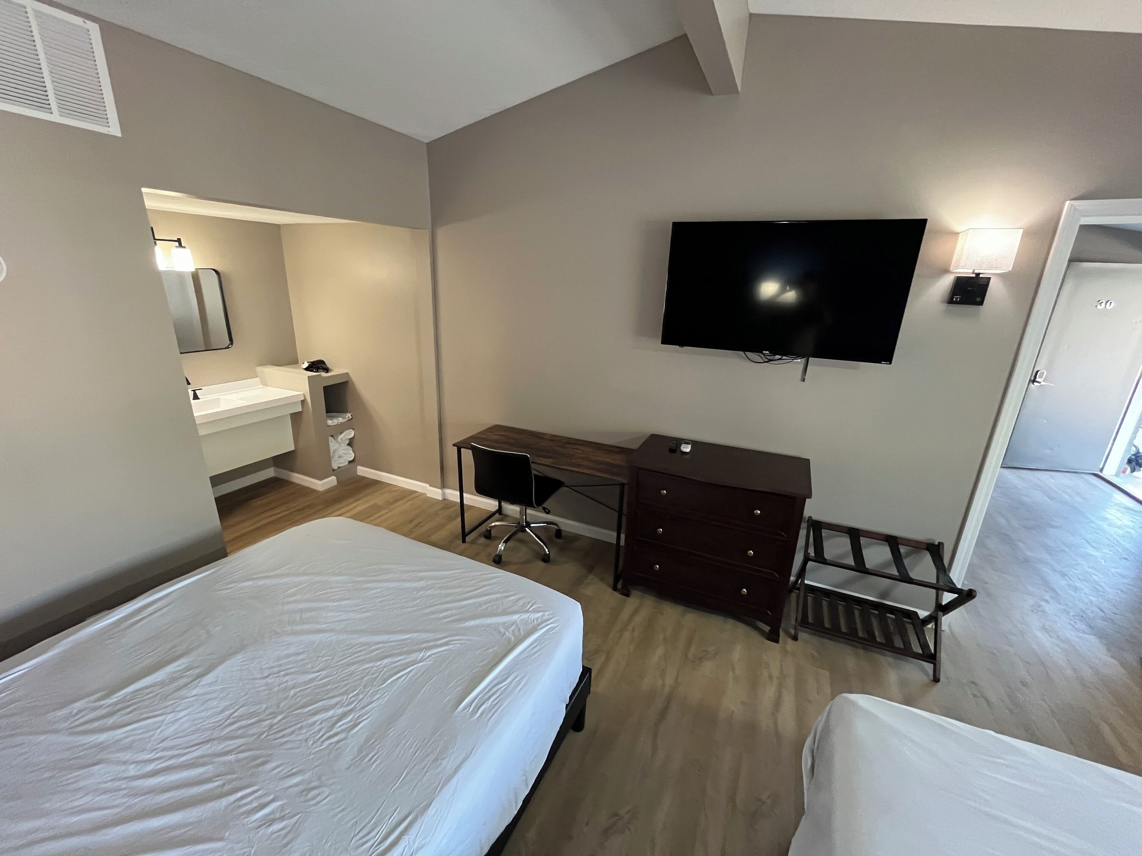 A hotel room with two beds, a desk, a TV, and a bathroom with a sink and mirror.