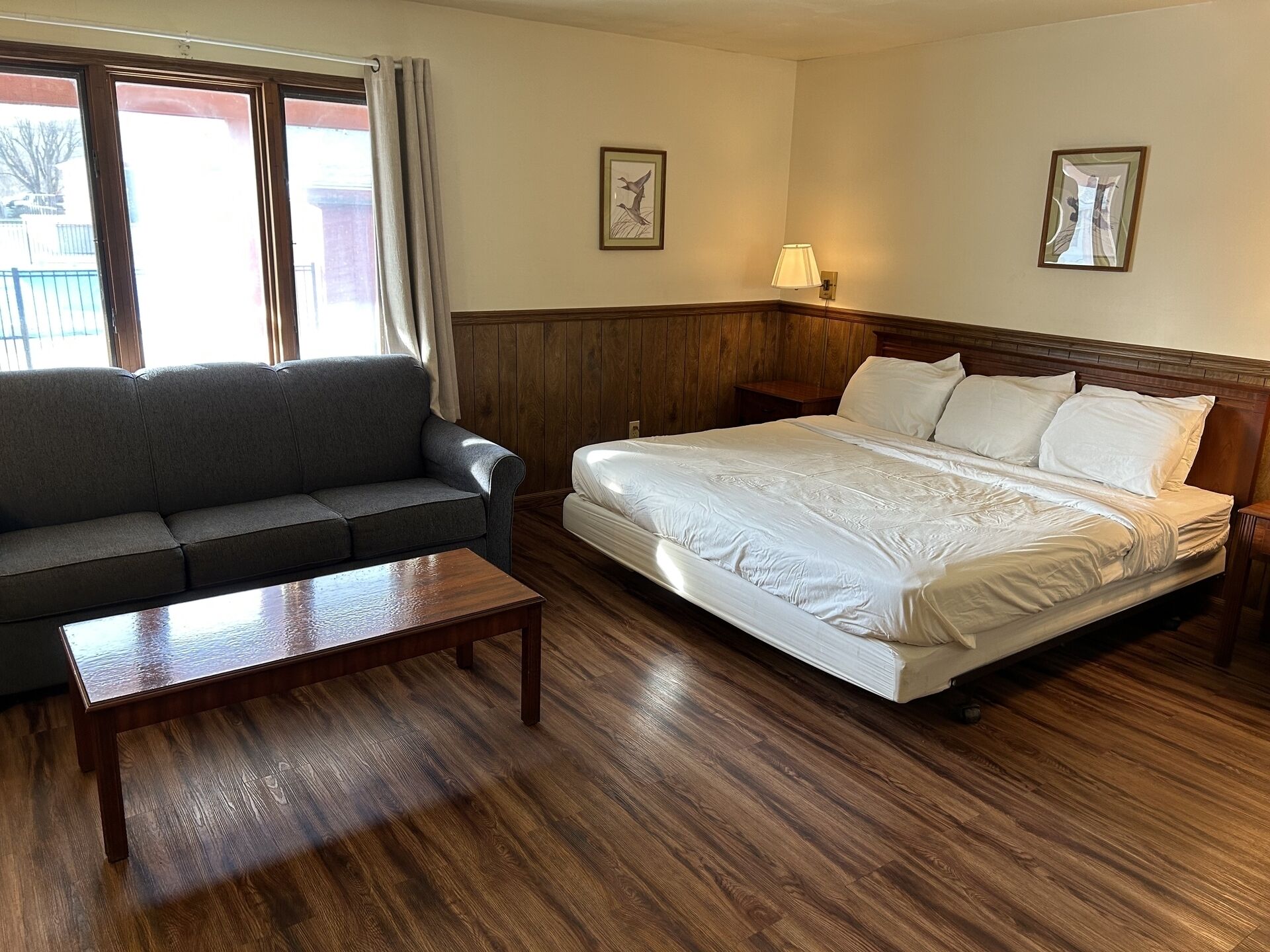 A hotel room with a wooden floor, a bed with white bedding, a gray sofa, a wooden nightstand, and a lamp.
