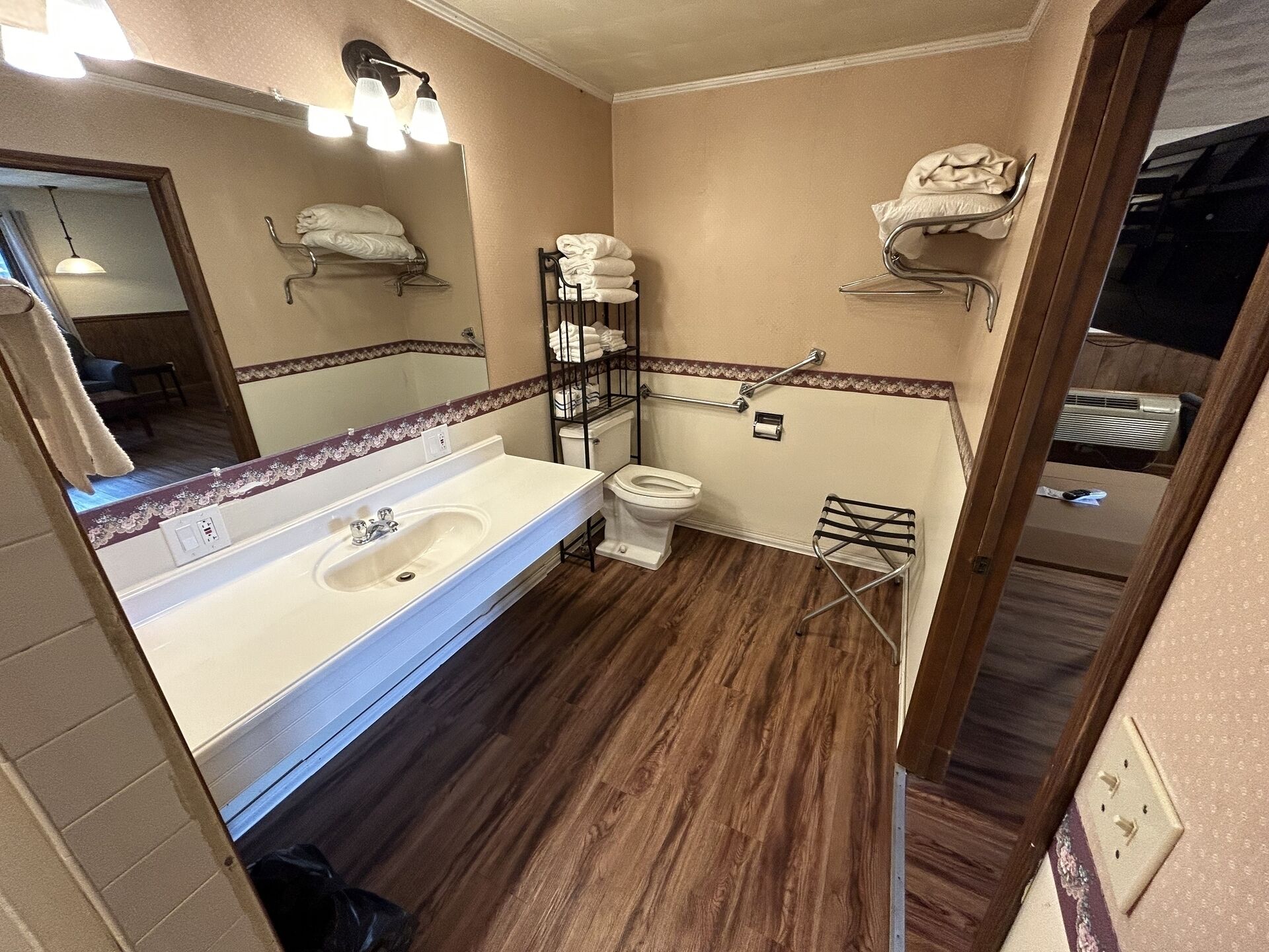 Une salle de bain avec un lavabo blanc, des toilettes et un miroir. On y trouve des porte-serviettes fixés aux murs et un plancher en bois.