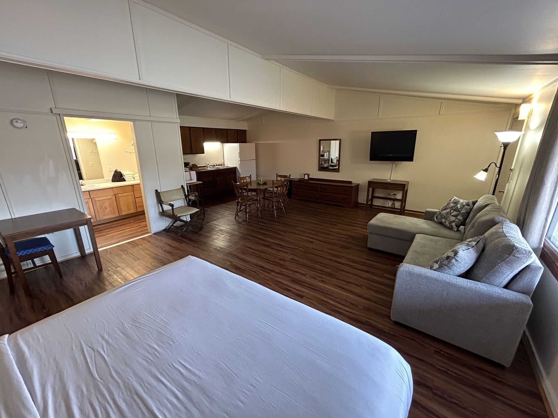 Une pièce spacieuse avec un grand lit, un canapé gris, un coin repas avec des chaises en bois, une cuisine avec des meubles blancs et un téléviseur à écran plat fixé au mur.
