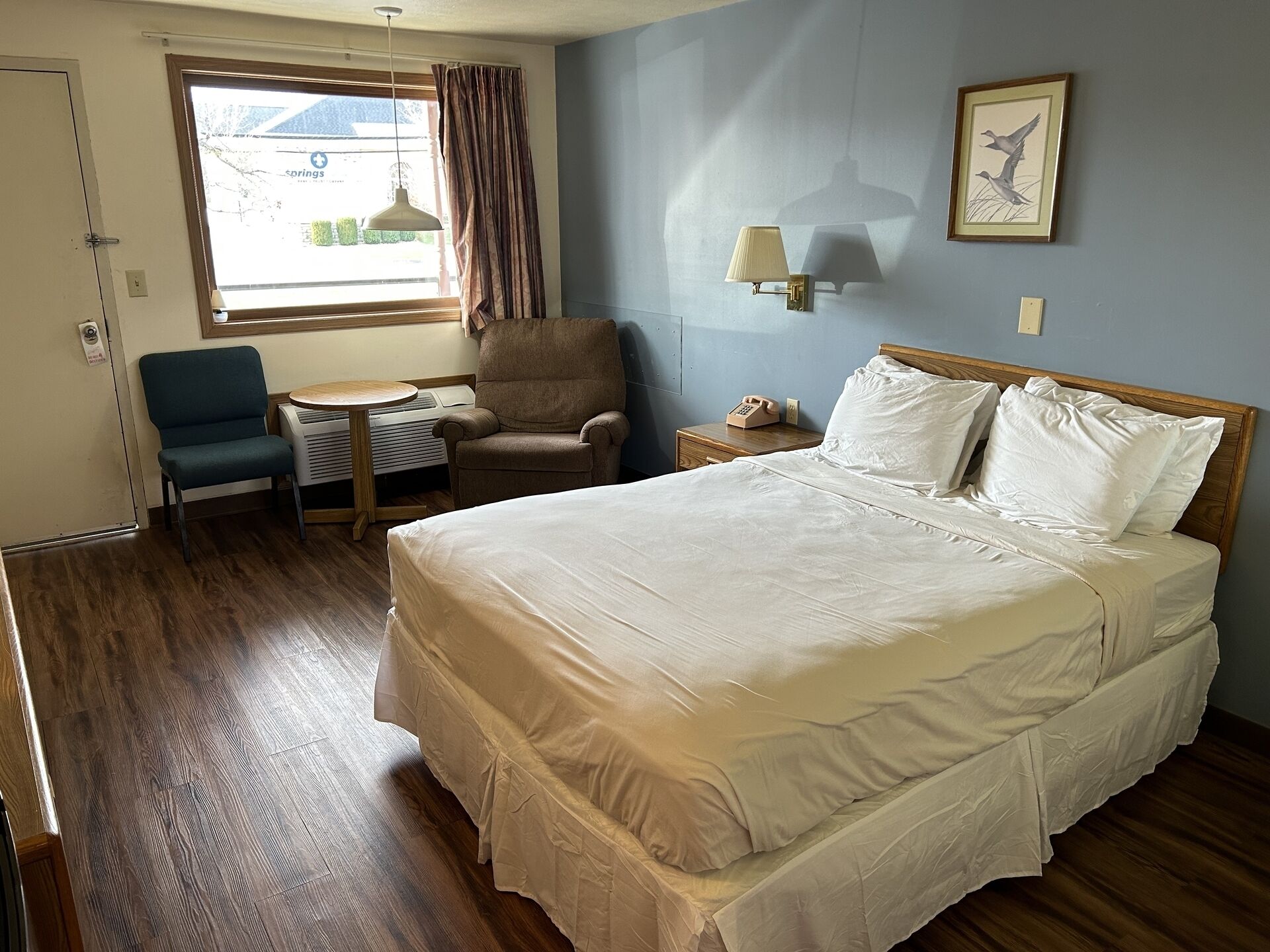 A hotel room with a bed, a chair, a small table, a lamp, and a window with curtains.