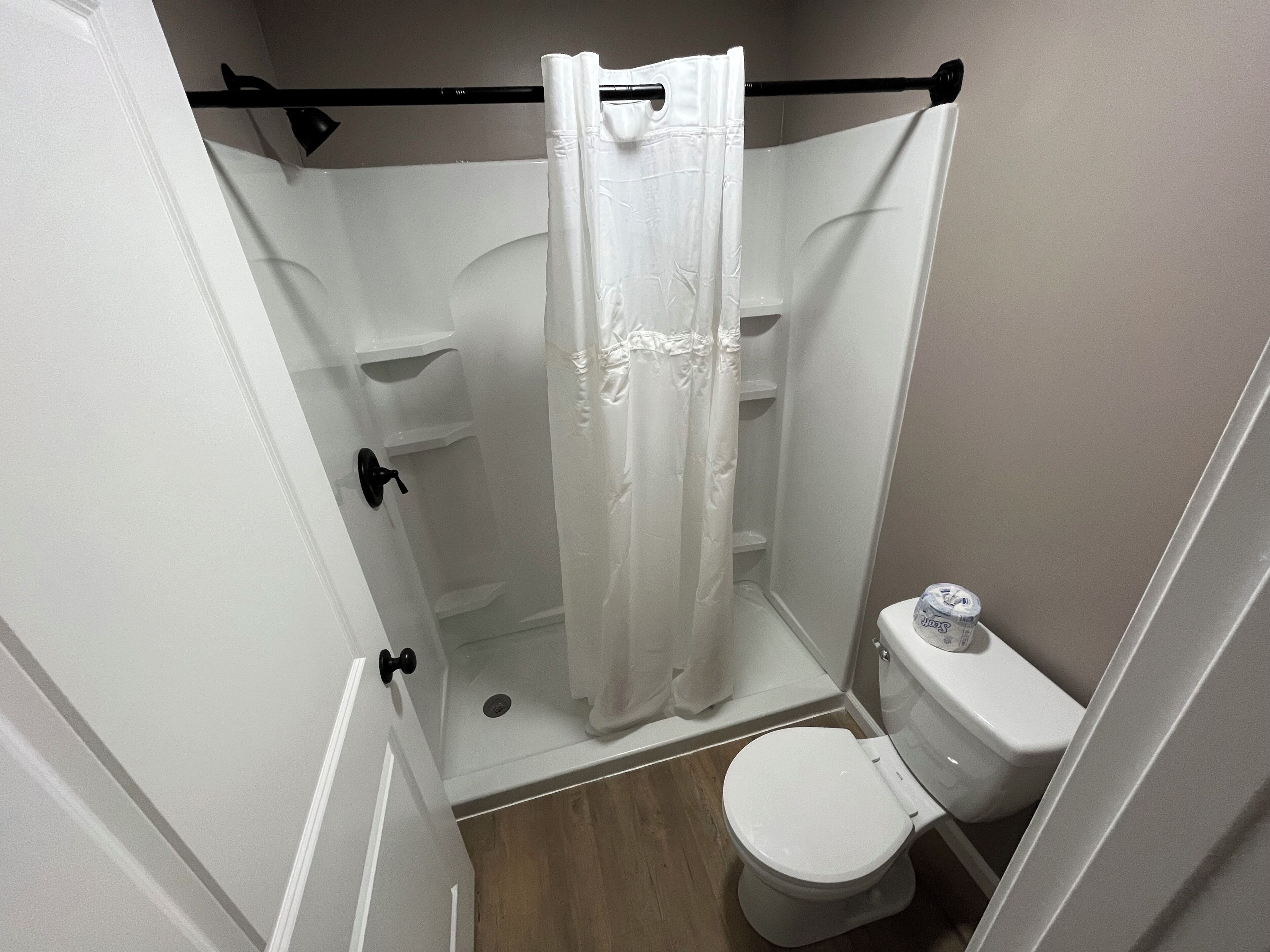 A small, enclosed shower area with a white shower curtain, a toilet, and a wooden floor.