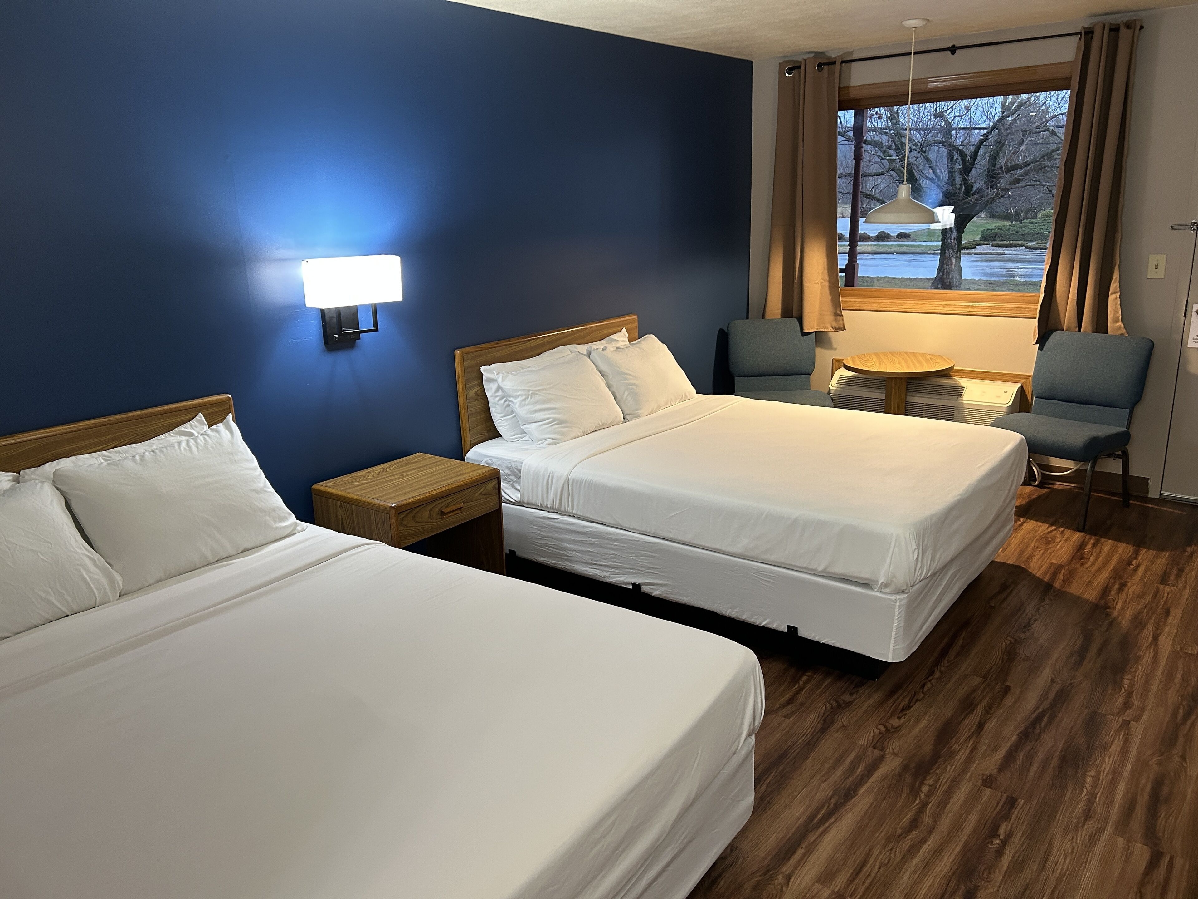 A hotel room with two beds, a nightstand, a chair, a small table, and a window with a view of a tree and a body of water.