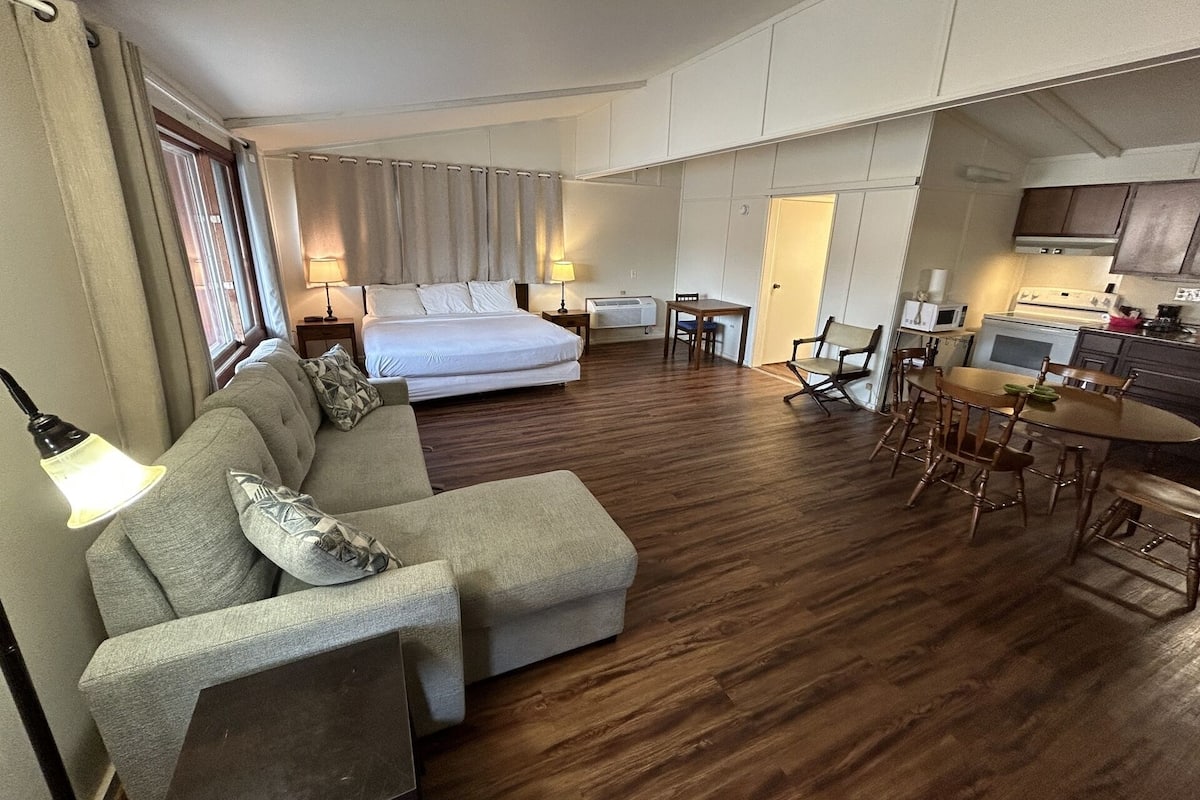 Ein Hotelzimmer mit Bett, einer Couch, einem Esstisch mit Stühlen, einer Küchenzeile und einem Fenster mit Vorhängen.