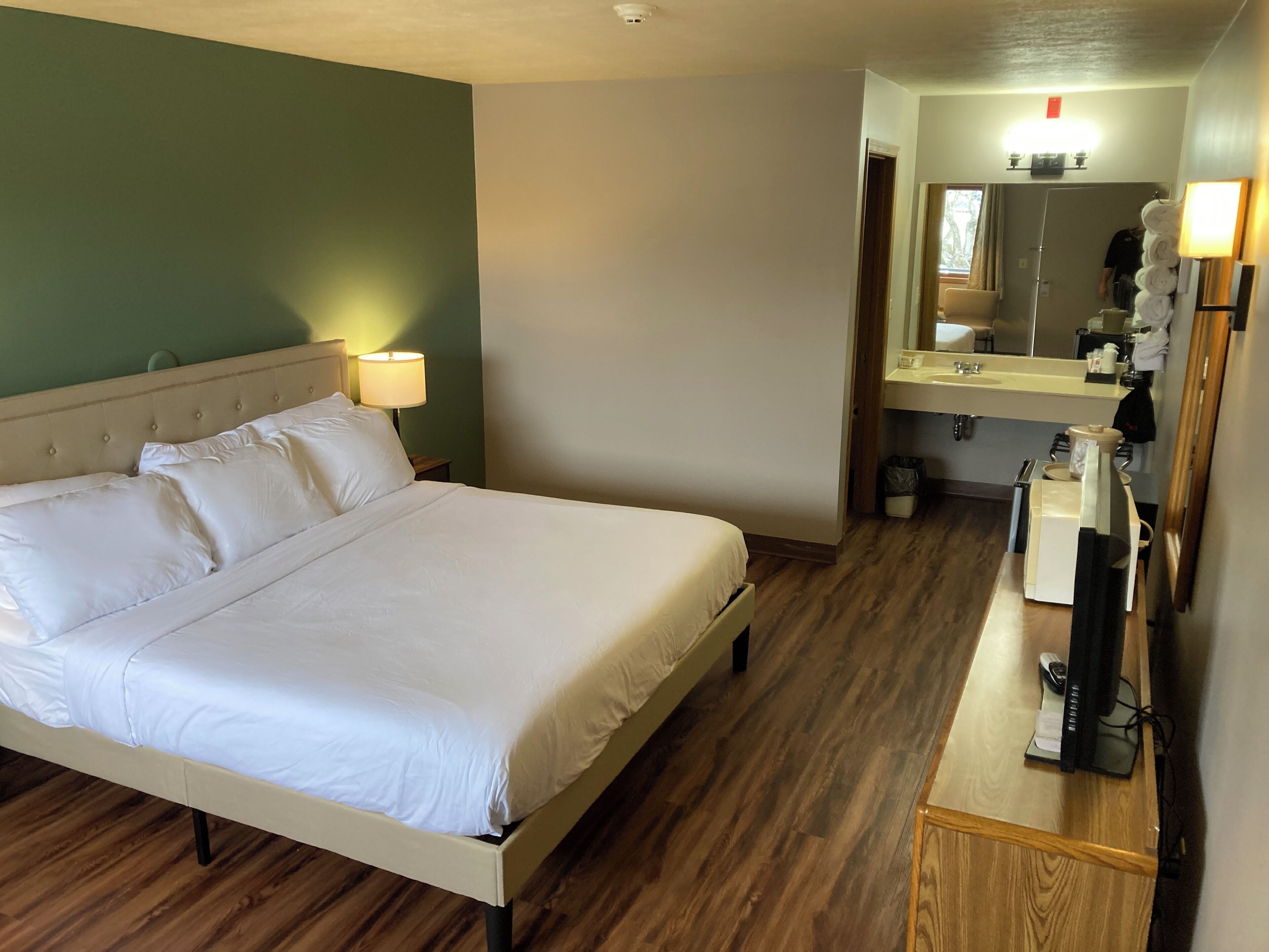 A hotel room with a bed, a nightstand with a lamp, a wooden desk with a telephone and a computer, a mirror, and a bathroom visible through an open door.