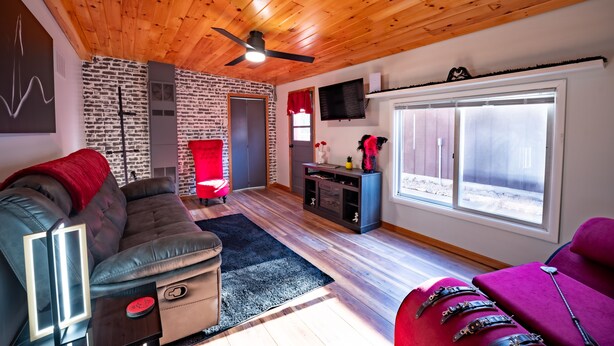 Un salón moderno con techo de madera, un sofá de cuero negro, un sillón rojo, un mueble azul y un ventanal con persianas.
