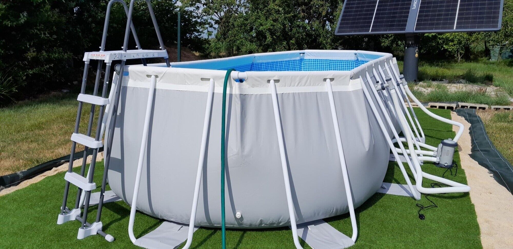 An above-ground swimming pool with a ladder, surrounded by artificial grass, and a solar panel in the background.