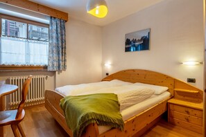 Een slaapkamer met een houten bed, een nachtkastje, een stoel, een raam met gordijnen, een radiator en een wandlamp.