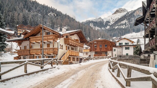 Un village enneigé, avec des bâtiments en bois, un chemin de terre et une chaîne de montagnes en arrière-plan.