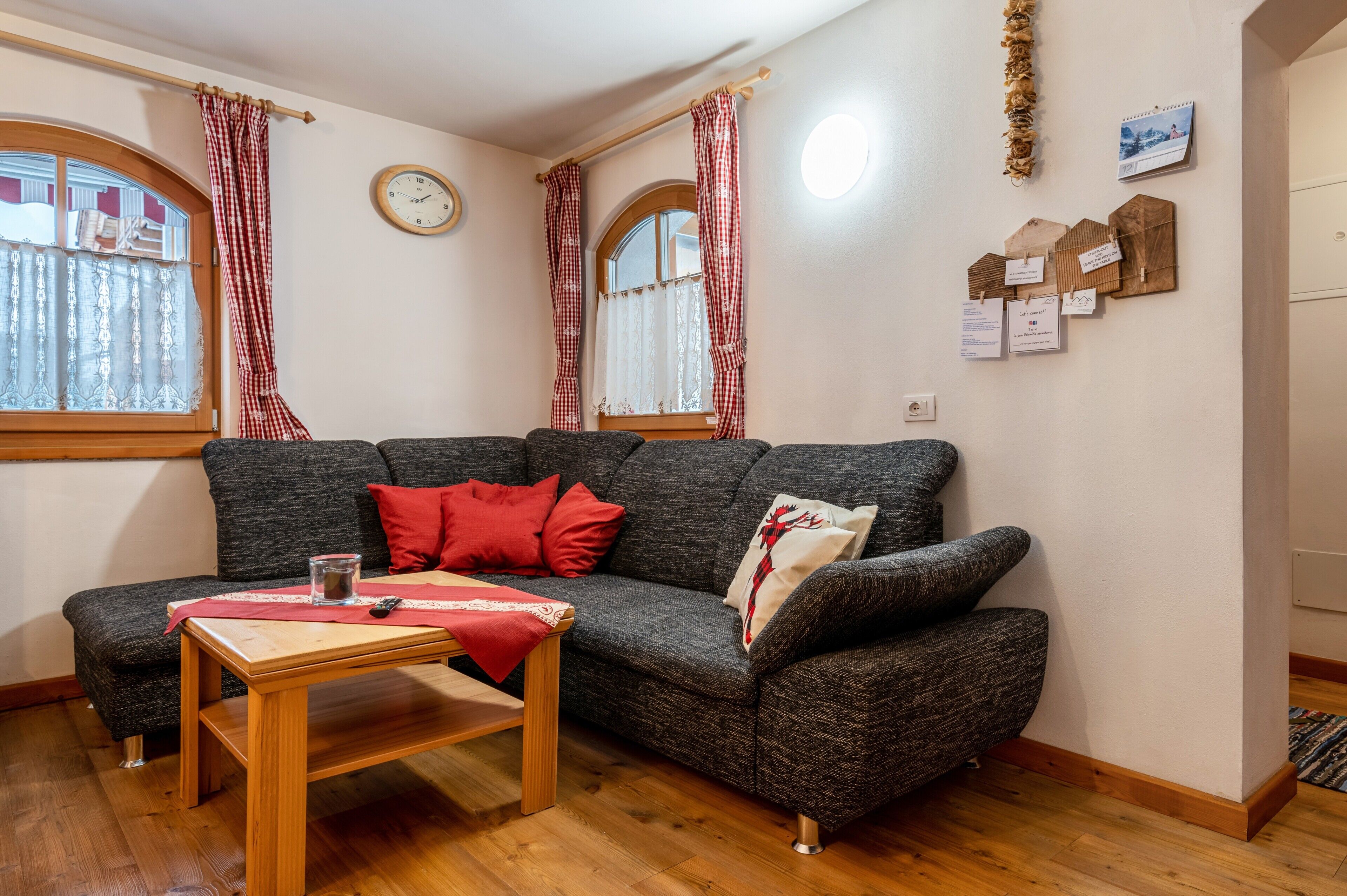 A cozy living room with a grey sofa, red cushions, a wooden coffee table, and a window with checkered curtains.