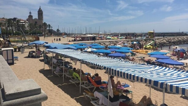 Ein Strand mit Reihen von gestreiften Strandsonnenschirmen und Liegestühlen, einer Steinmauer und einem in der Ferne liegenden Gebäude mit einem Glockenturm.
