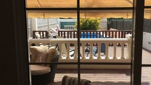 Blick durch ein Fenster auf eine Holzdielen-Terrasse mit weißer Reling, ein Schwimmbad und Gartenmöbel.