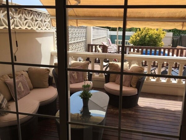 A balcony with wicker furniture, a glass table, and a view of the pool and greenery.