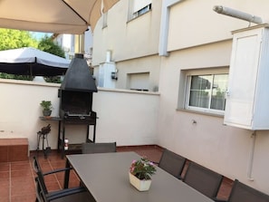 Une terrasse extérieure avec un barbecue, une table, des chaises et un pot de fleurs.