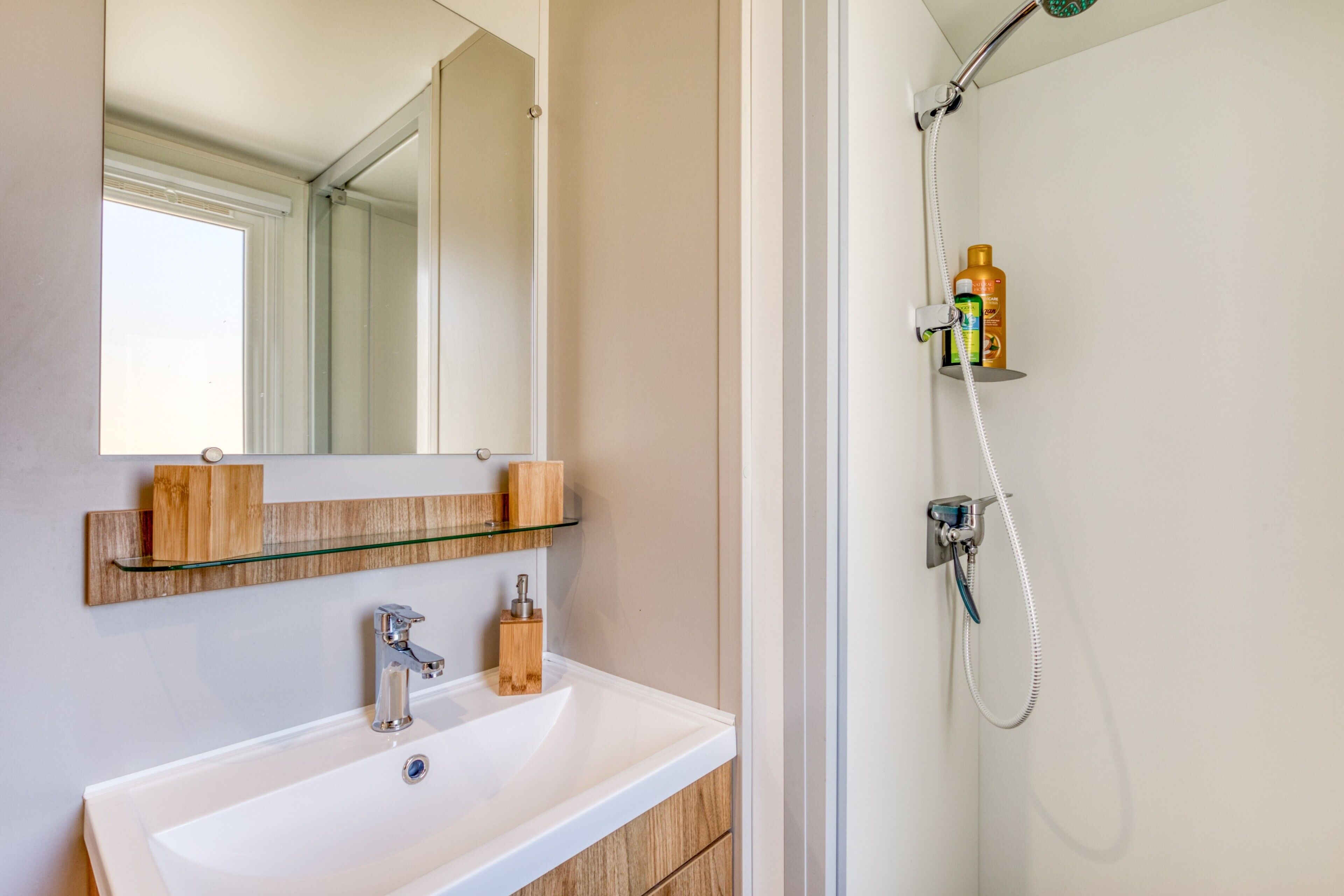 A bathroom with a white sink, a mirror, a glass shelf with toiletries, and a shower with a handheld showerhead.