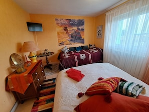 Quarto com cama, televisão, mesa com abajur, pintura na parede e janela com cortinas.