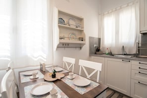 A dining area with a table set for four, white chairs, a kitchen with a sink and cabinets, and a shelf with decorative plates. - Holiday Home 'Punda Beach Seaside Retreat' with Sea View, Wi-Fi and Air Conditioning (Paros)