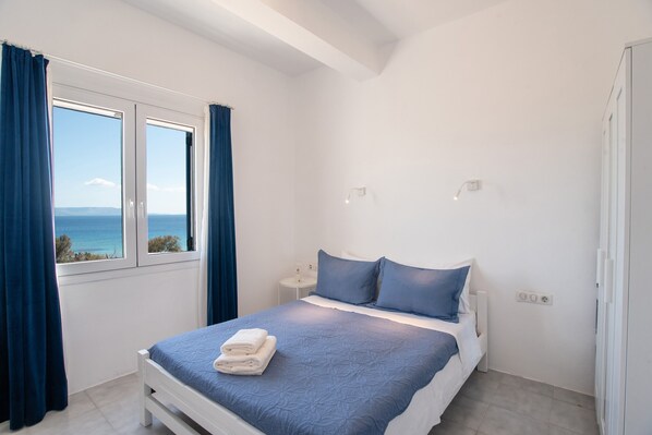 Une chambre à coucher avec un lit, une fenêtre avec des rideaux, une table de chevet et une vue sur la mer.