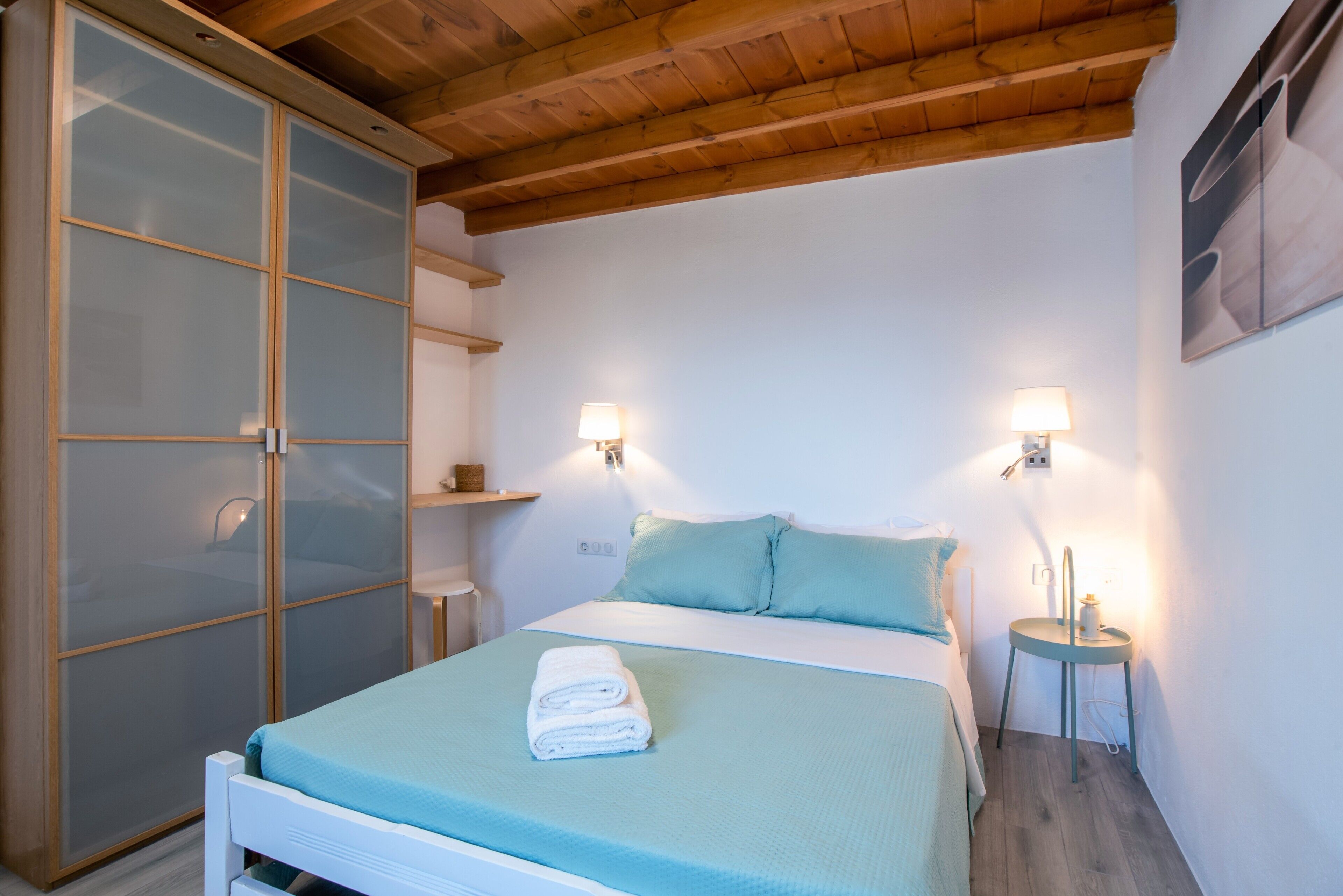 A bedroom with a bed, two wall-mounted lamps, a wardrobe with frosted glass doors, and a wooden ceiling.