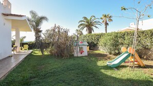 Ein grünes Areal im Freien mit einer kleinen Spielhütte, einer Rutsche und einigen Stühlen.