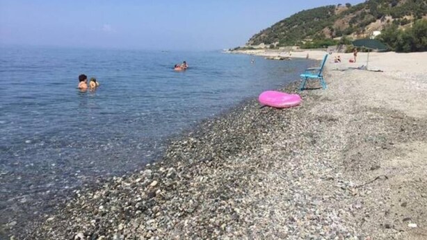 Una playa de canto rodado con agua cristalina, algunas personas nadando y un flotador rosa en la orilla.