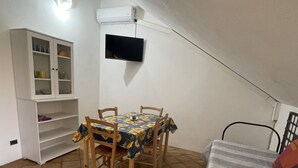 Pequeña habitación cerrada con suelo de baldosas, una mesa de comedor con sillas, un sofá, un armario y un televisor de pared.
