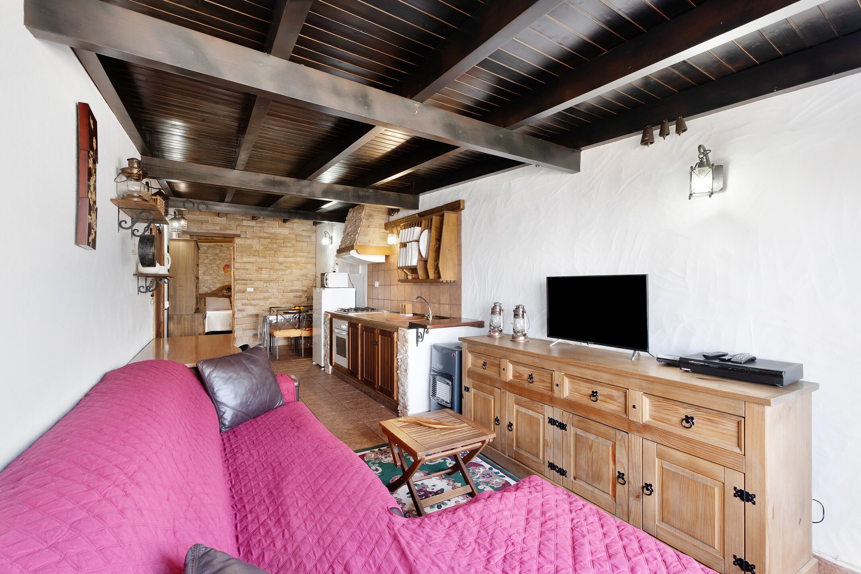 A living room with a pink sofa, wooden cabinets, a TV, and a kitchen area with a brick wall and wooden ceiling beams.