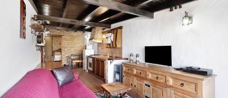 A living room with a pink sofa, wooden cabinets, a TV, and a kitchen area with a brick wall and wooden ceiling beams.