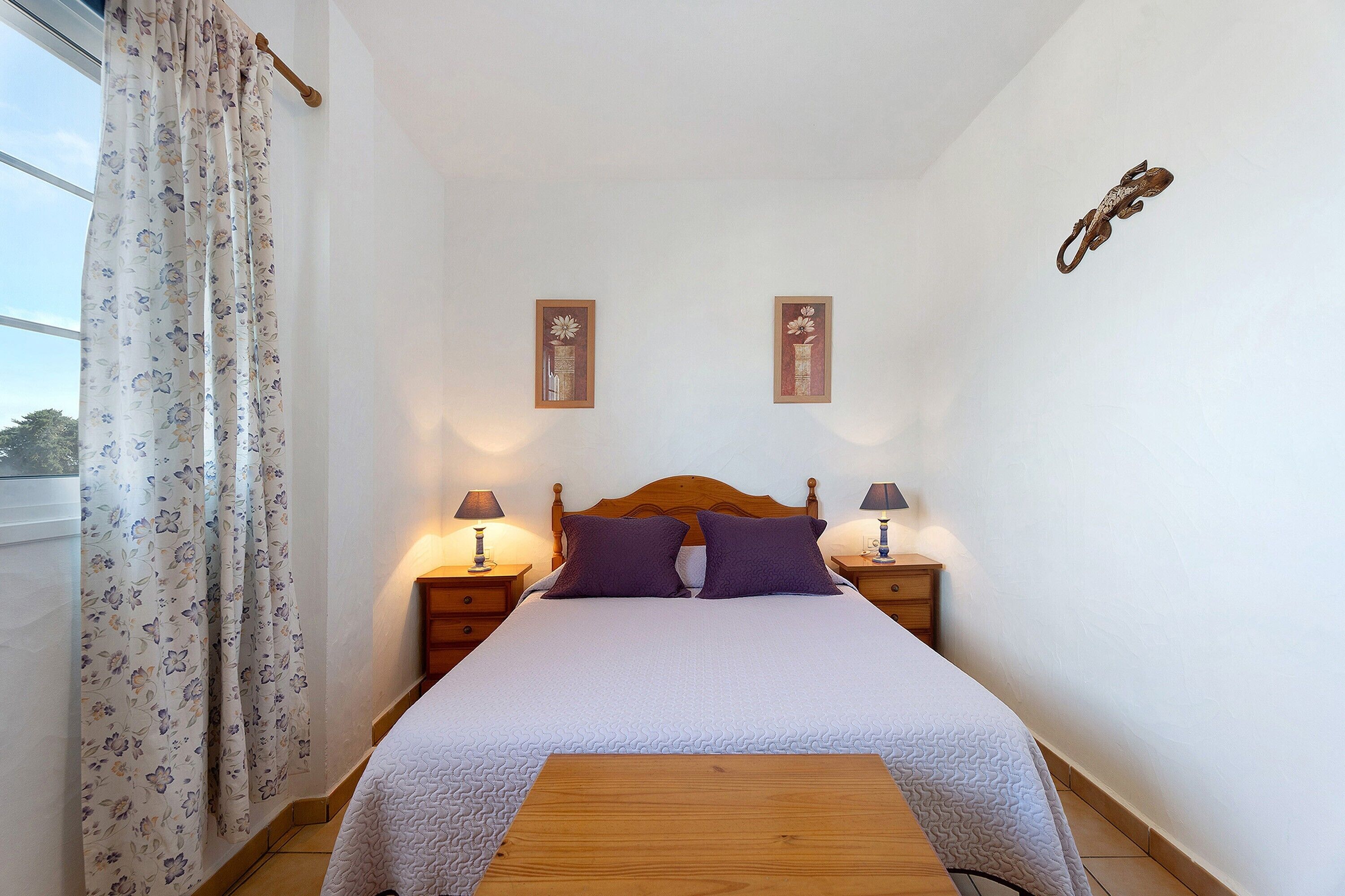 Een net opgemaakt bed met een houten hoofdbord, twee nachtkastjes met lampen, en een raam met bloemen gordijnen.