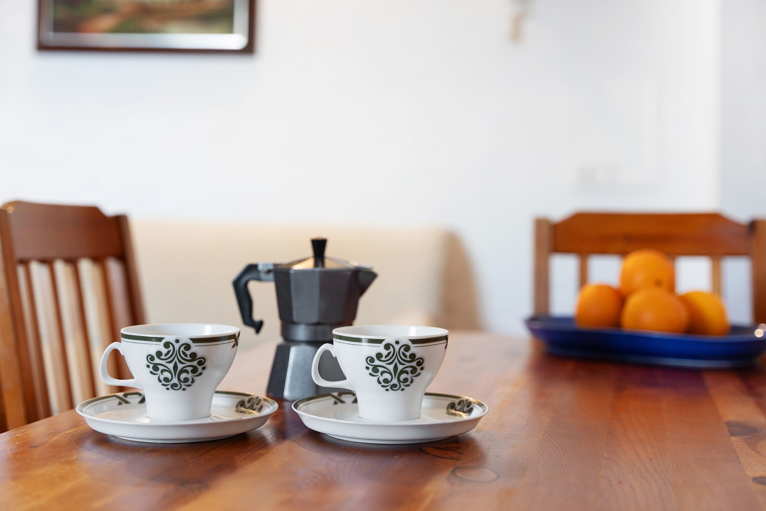 Dos tazas blancas con diseños verdes sobre una mesa de madera, una cafetera y un plato azul con naranjas al fondo.