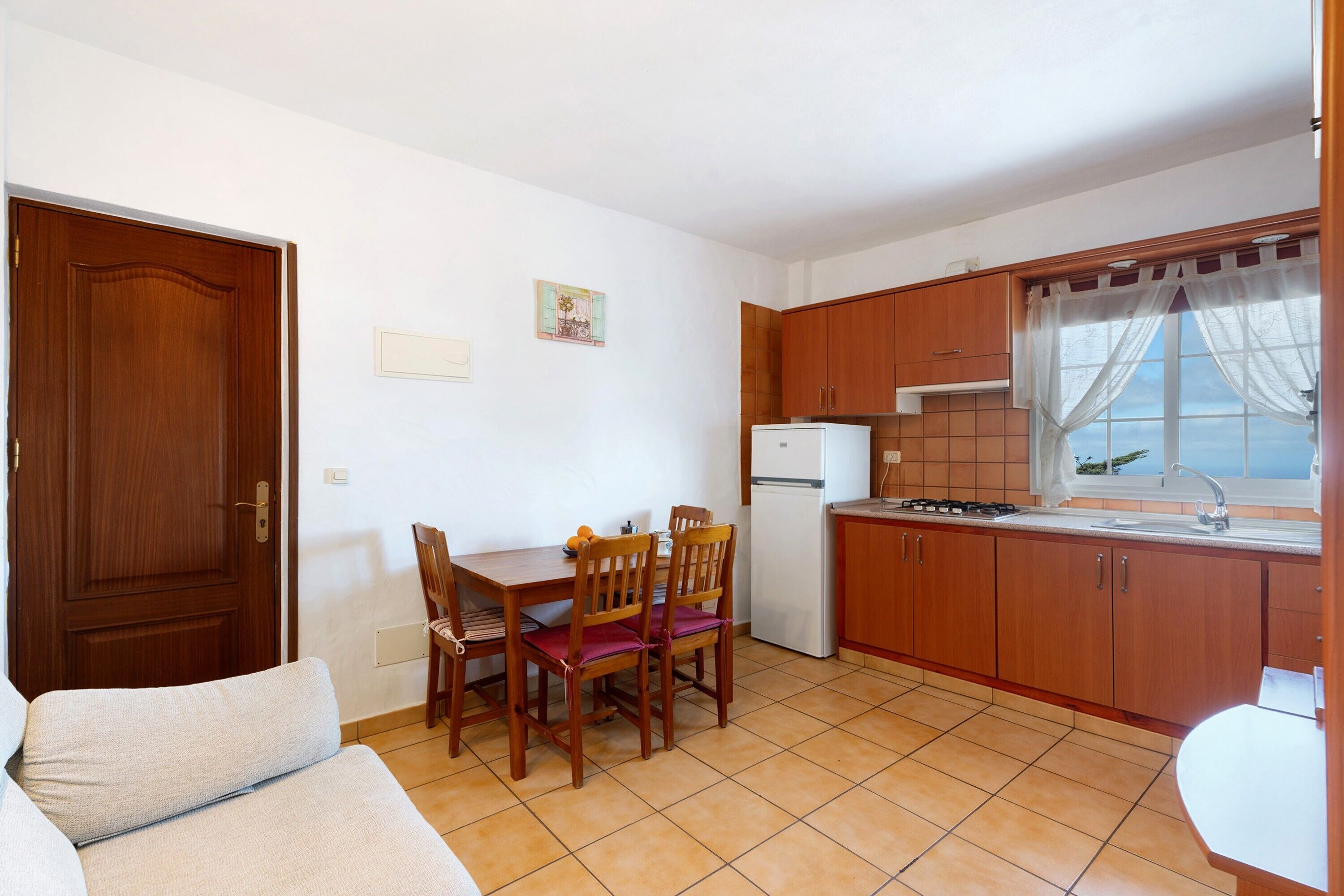 A compact kitchen with wooden cabinets, a dining table with chairs, a refrigerator, and a window with curtains.