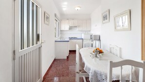 Une cuisine étroite avec des meubles blancs, une table à manger avec des chaises et une fenêtre avec des stores.