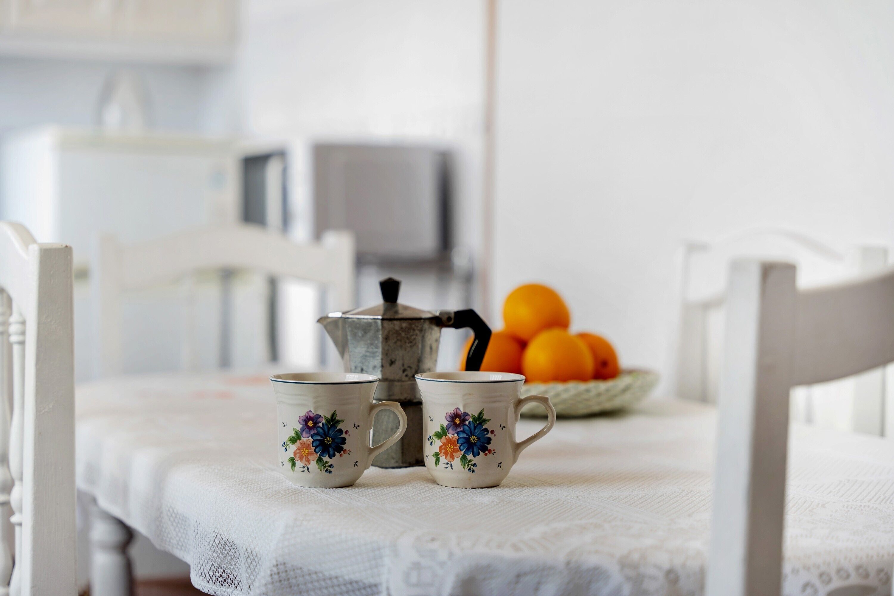 A table with two floral mugs and a coffee pot, a bowl of oranges, and a white chair.