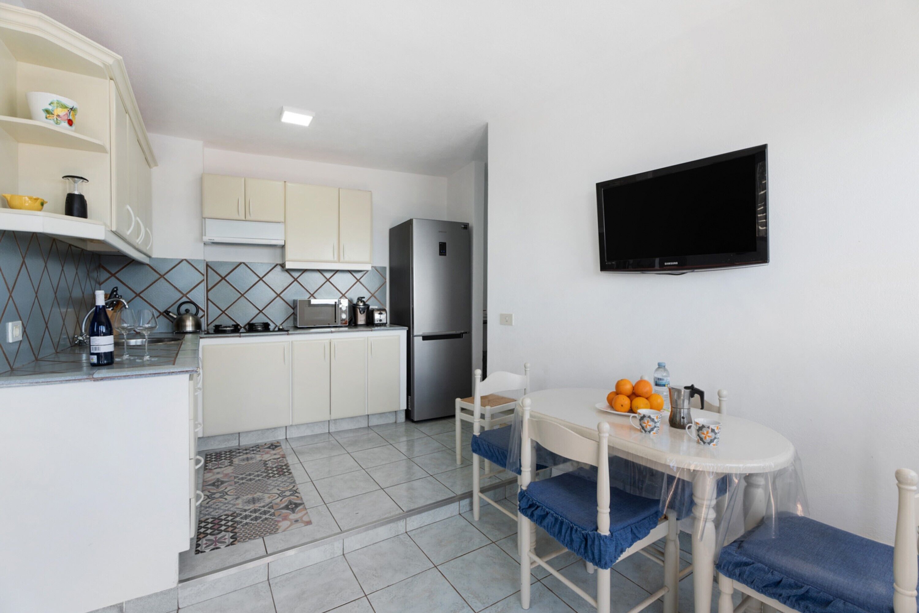 A compact kitchen with white cabinets, a refrigerator, a dining table with blue chairs, and a mounted TV.