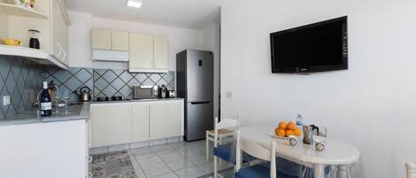 A compact kitchen with white cabinets, a refrigerator, a dining table with blue chairs, and a mounted TV.