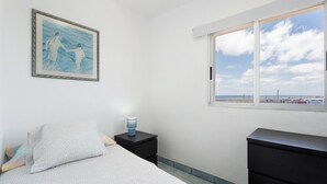 Une chambre à coucher comprenant un lit, une table de chevet, une commode, une fenêtre donnant sur la mer et un tableau accroché au mur.