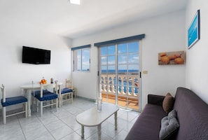 Quarto com televisão, mesa, cadeiras, sofá e varanda com vista.