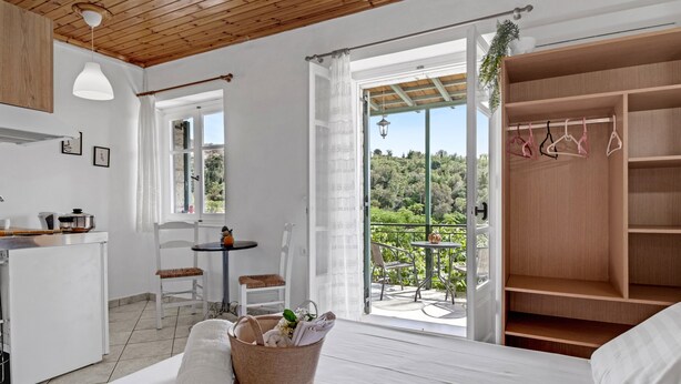 A bedroom with a bed, a small table, chairs, a kitchen area, and a balcony with a view of greenery. - Studio Apartment 'With Sea View' with Sea View, Wi-Fi and Air Conditioning (Drimonas)