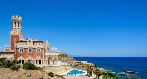 Un edificio histórico con una torre, piscina y vista al mar.