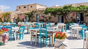 Un café en plein air, avec des tables et des chaises, entouré de bâtiments en pierre aux portes bleues.
