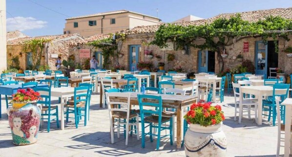 An outdoor café with tables and chairs, surrounded by stone buildings with blue doors.