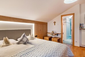 Un dormitorio con cama, mesitas de noche, cabecero de madera, una cruz en la pared y una puerta que da al baño.