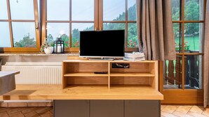 A room with a TV on a wooden stand, a window with curtains, and a view of greenery outside. - Apartment 'Zunerhof Abendsonne' with Mountain View, Balcony and Wi-Fi (Lengstein Ritten)