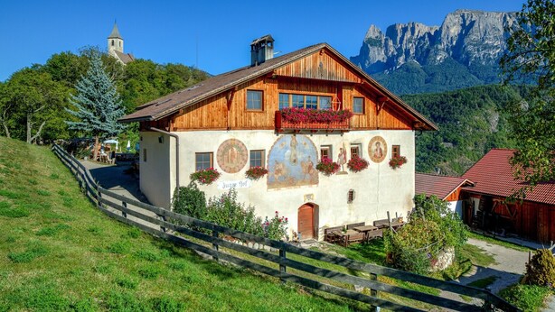 A wooden house with a balcony, a church in the background, and mountains. - Apartment 'Zunerhof Abendsonne' with Mountain View, Balcony and Wi-Fi (Lengstein Ritten)