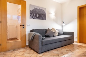 Una habitación con un sofá gris, una puerta de madera, una lámpara de pared y un cuadro enmarcado.